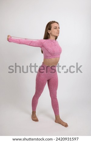 A girl athlete in a pink training suit shows warm-up exercises with a stretching elastic band. Close-up on a light background.