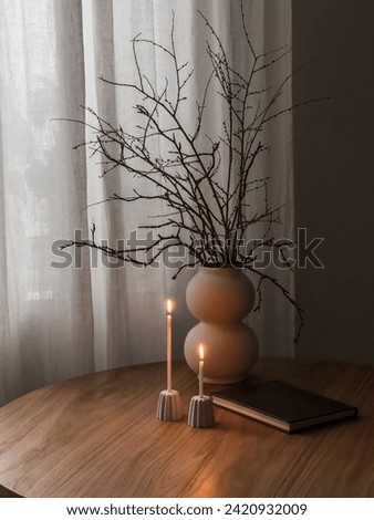 Cozy evening interior - thin candles in ceramic candlesticks, a vase with a bouquet of dry branches, a book on a wooden round table in the living room Royalty-Free Stock Photo #2420932009