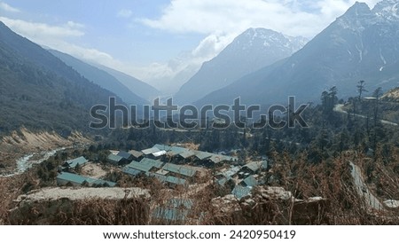 The image shows a serene and picturesque landscape of a mountainous region. A small settlement with green roofs is nestled amidst the rugged terrain, surrounded by towering mountains veiled in mist.