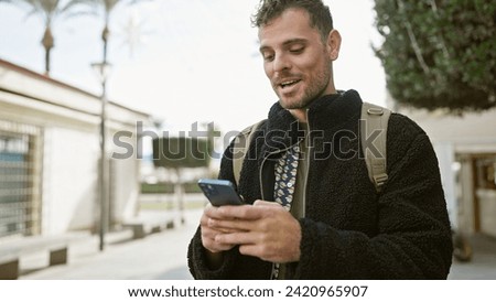 Smiling bearded young man using smartphone on a sunny city street, depicting urban life and technology. Royalty-Free Stock Photo #2420965907