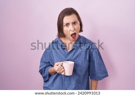 Middle age hispanic woman drinking a cup coffee in shock face, looking skeptical and sarcastic, surprised with open mouth  Royalty-Free Stock Photo #2420991013