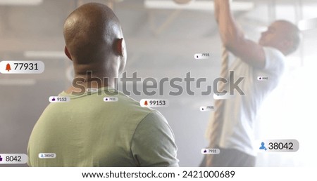 Image of social media notifications over diverse male trainer and man climbing rope at gym. Fitness, exercise, strength, communication, digital interface and technology digitally generated image.