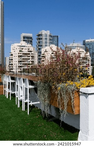 Urban rooftop garden with vibrant flowers and white chairs, overlooking modern cityscape under clear blue sky in Ramat Gan, Israel