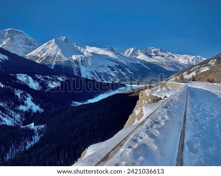 A picture taken of a mountain on the roadside in Alberta, Canada. There is a forest of pine trees on the mountain and roadside. There is a wall and footprints in the snow. Sky bright with clouds. 