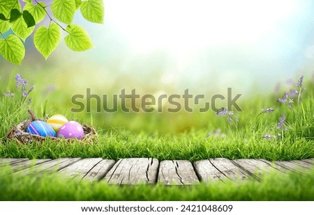 Painted easter eggs in a birds nest celebrating a Happy Easter in spring with a green grass meadow, tree leaves, bluebells and bright sunlight background and rustic wooden bench to display products. Royalty-Free Stock Photo #2421048609