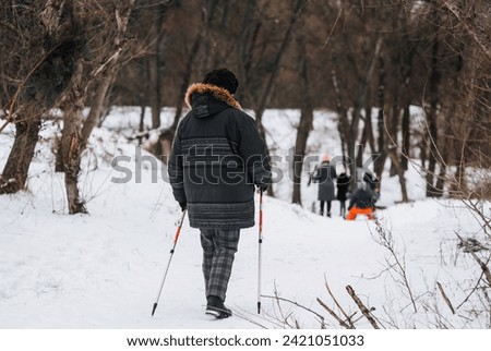 An adult, elderly woman with aluminum hiking sticks walks along the road in winter on white snow on the street outdoors. Photography, healthy lifestyle concept.