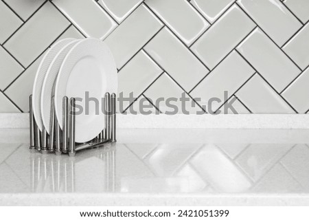 A stack of clean white plates in a plate rack on a reflective kitchen countertop, with a tiled kitchen background, copy space Royalty-Free Stock Photo #2421051399