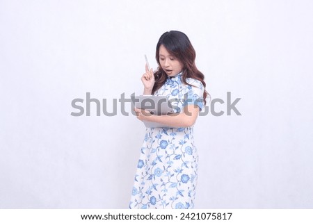 Beautiful shocked Indonesian Chinese woman wearing modern Chinese blue dress carrying tablet and looking at it isolated white background