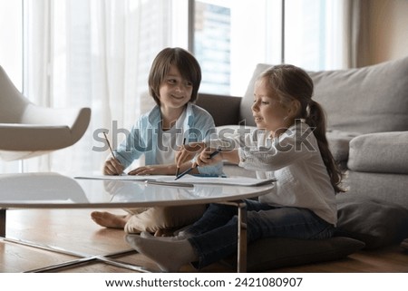 Two little beautiful boy and girl, friends, cousins spend pastime together sit at table in cozy, warm living room drawing pictures in paper album with colored pencils enjoy leisure and friendly talk