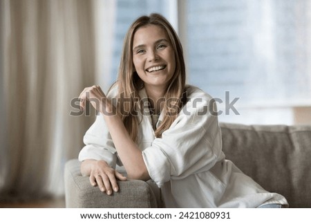 Happy beautiful woman relaxing alone on cozy sofa in modern living room smile staring at camera, enjoy carefree pastime in rented or own fashionable apartment. Tenancy, independence, untroubled mood