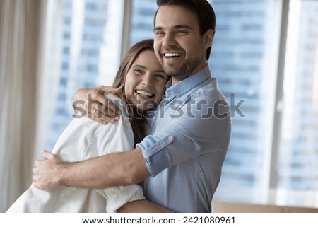Close up portrait beautiful millennial spouses laughing, cuddling standing in modern apartment with city skyscrapers view. Homeowner family, bank loan, tenancy, start new life, cohabitation, marriage Royalty-Free Stock Photo #2421080961