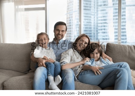 Young wife and husband posing for family photo sit on sofa with two little kids, smile look at camera, spend time together at modern apartment. Bank loan, new dwelling owners portrait, custody, love Royalty-Free Stock Photo #2421081007