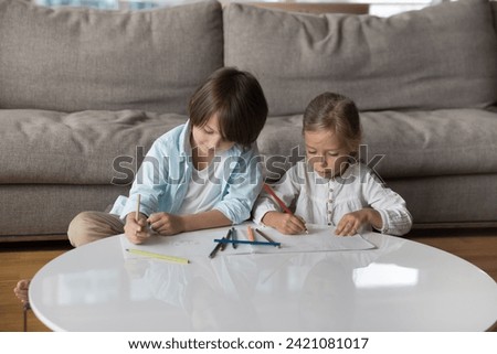 Two focused cute little children spend weekend in living room, sit on warm floor at table, draw pictures in sketchbook with colored pencils, brother and sister enjoy creative leisure at home. Hobby