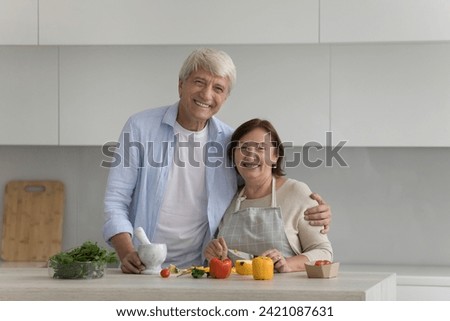 Happy senior retired family couple home portrait. Cheerful older husband and mature wife enjoying culinary hobby together, cooking salad from fresh vegetables in kitchen, hugging, laughing
