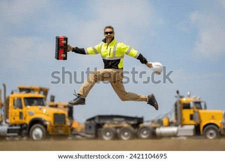 Middle aged builder excited jump on site construction. Excited builder construction worker in a safety helmet jumping in front of the trucks. Excited crazy builder man in helmet jump outdoor. Royalty-Free Stock Photo #2421104695