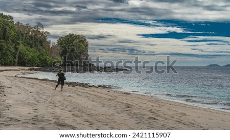 A girl stands on a sandy beach, taking pictures of a picturesque rock. The view from the back. Footprints in the sand. A calm turquoise ocean. Clouds in the blue sky. Madagascar. Nosy Tanikely 
