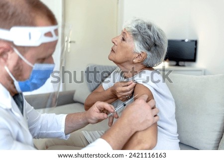 Nurse with protective face mask on, at home giving injection to an old woman during corona outbreak. Male Nurse applying vaccine on female senior patient's arm in his bright office during the day.