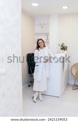 Confident female aesthetician in white coat standing in a modern clinic with minimal decor, welcoming with a warm smile. Royalty-Free Stock Photo #2421159053