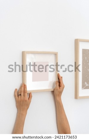 Cropped shot of woman holding wooden picture frame in hands. Vertical shot of female with copy space art placard against wall. Small square mockup of art painting in white minimalist room