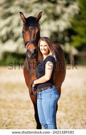 Young woman with a black shirt and short highlighted hair stands with her horse on a sunny riding arena.