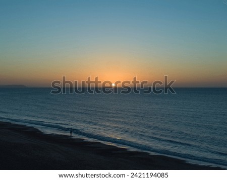 A lone male walks along the tideline at dawn on a Winters morning. There are no clouds in the January blue and orange sunrise sky and the sea is calm.