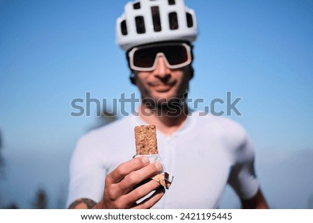Male cyclist wearing cycling kit and helmet is holding energy snack for endurance during bicycle training. Sport nutrition. Sport snack for cyclist. Granola energy bar. Royalty-Free Stock Photo #2421195445