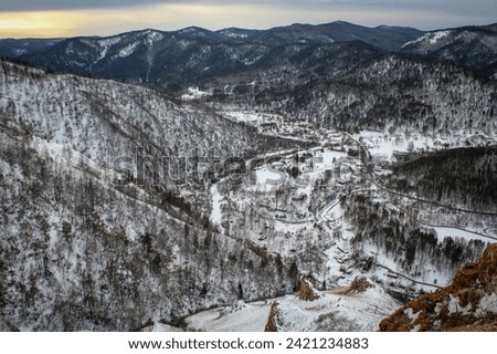 Nature froze in anticipation of spring. Winter gloomy Natural landscape at dusk. Village between hillsides, view from hill. Road, frozen river and buildings. Hiking area near home.  Royalty-Free Stock Photo #2421234883