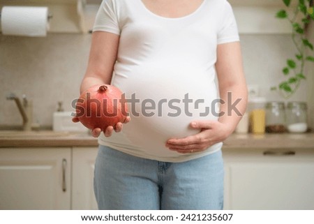 A pregnant woman holds a pomegranate fruit in her hands while standing in a home kitchen. Pregnancy and proper nutrition while expecting a child