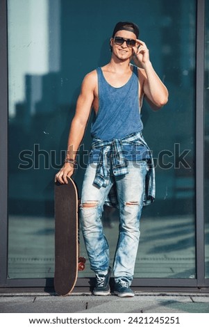 Stylish guy in casual clothes, cap and glasses is holding a skateboard and smiling while walking in the city