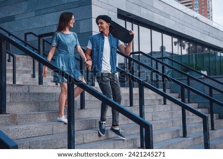 Stylish young couple is holding hands and smiling while walking outdoors, guy is holding a skateboard
