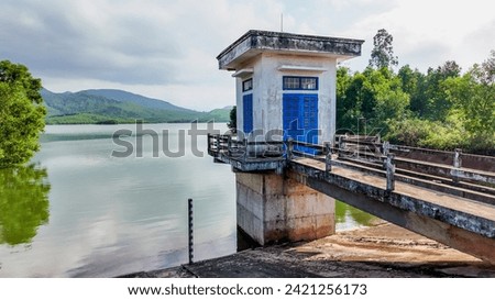 A small blue doored water pump house on a concrete platform extends over a tranquil lake with lush green hills in the background Royalty-Free Stock Photo #2421256173