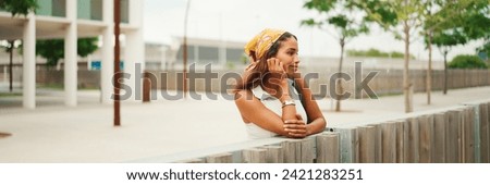 Profile of cute tanned woman with long brown hair wearing white top and yellow bandana stands thoughtfully leaning against the railing and looking into the distance cityscape background, Panorama