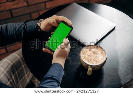 Man sitting and holding smartphone with green screen in cafe with coffee latte and laptop. Freelancer and remote work