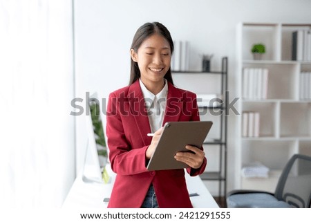 Professional young Asian businesswoman smiling while working on a digital tablet in a modern office setting. Royalty-Free Stock Photo #2421291555