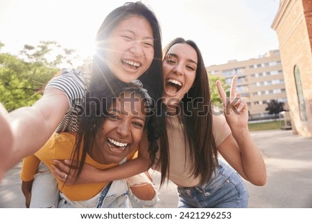 Three young multiracial friends taking selfie portrait together outdoors in summer. Latin woman giving piggyback ride to Asian female on city street. Excited girls posing for funny photo, lens flare.