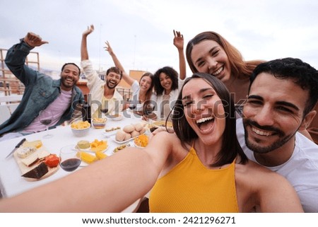 Excited young pretty woman taking group selfie with joyful friends on rooftop. Cheerful people poses making smiling faces to camera outdoor gathered sitting table at lunch raising happy arms for photo