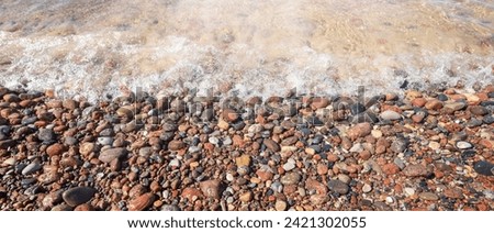 Panoramic photo of sea coast with brownish red pebbles and shining sea wave.