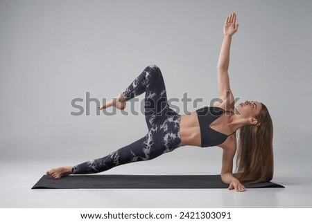 Caucasian female athlete in dark activewear doing side plank with bent leg in studio. Side view of concentrated sportswoman practicing variation of side plank asana, isolated on grey. Yoga concept. Royalty-Free Stock Photo #2421303091