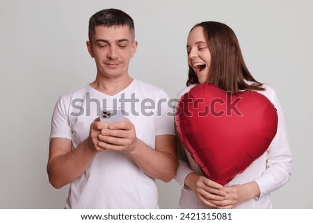 Man and laughing woman wearing white clothing holding heart shaped air balloon isolated over gray background using cell phone browsing internet web pages