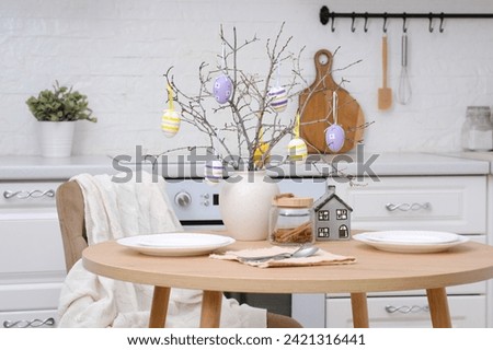 Home spring easter design in kitchen. Plate with cookies on round wooden table, creamic vase with spring branches with easter eggs, beige chair with cover in white kitchen. High quality photo.