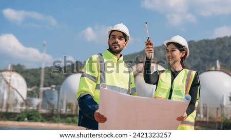 Two engineers in safety gear with blueprints discussing work in front of industrial storage tanks on a clear day.