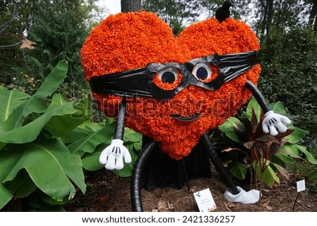 This image captures a whimsical, smiling heart-shaped character, bespectacled and sporting vibrant autumn flowers, creating a playful atmosphere in a garden setting. Royalty-Free Stock Photo #2421336257
