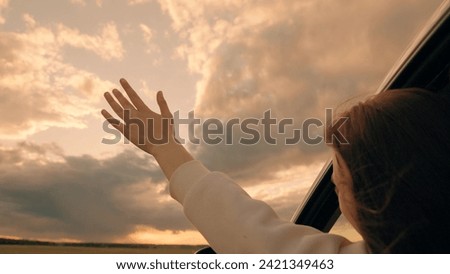 Girl waves her hand from car window, travels, catches wind with her fingers, Clouds. Road trip on way to vacation. Young woman driver plays, catches fresh wind with her hand from car window. Travel Royalty-Free Stock Photo #2421349463