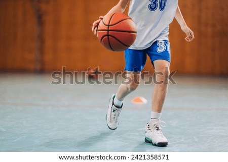 An unrecognizable junior basketball team member in action is dribbling a ball on court during training. Cropped picture of junior basketball player running and dribbling a ball on training on court.