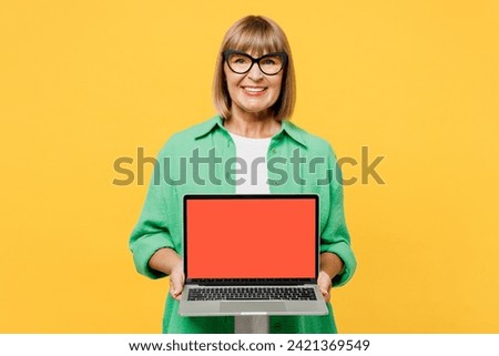 Elderly blonde IT woman 50s year old wear green shirt glasses casual clothes hold use work on laptop pc computer with blank screen workspace area isolated on plain yellow background. Lifestyle concept