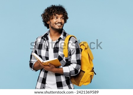 Young smiling Indian boy student he wears shirt casual clothes backpack bag read book hold notebook look aside isolated on plain pastel light blue background. High school university college concept