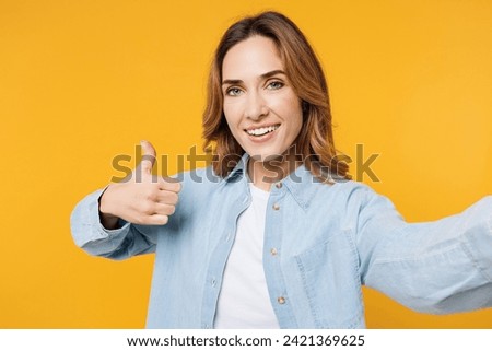 Close up young woman wear blue shirt white t-shirt casual clothes doing selfie shot pov on mobile cell phone show thumb up like isolated on plain yellow background studio portrait. Lifestyle concept