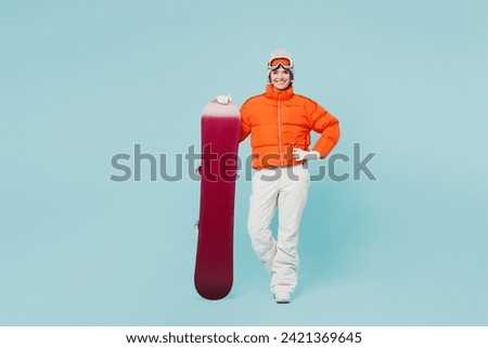 Full body happy young woman wear warm padded windbreaker jacket hat ski goggles mask hold snowboard look camera travel rest spend weekend winter season in mountains isolated on plain blue background