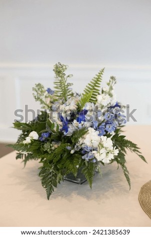 Floral centerpiece with blue and white stock flowers and green ferns. On table with white table cloth. 