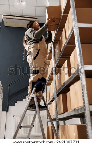 African american employee standing on ladder in storehouse, preparing customers orders using cardboard boxes. Small business worker wearing overall doing merchandise quality control in warehouse